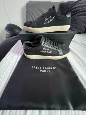 Saint Laurent svarta sneakers - Svarta sneakers från Saint Laurent med vit logga på sidan och svart läderdetalj. Klassisk låg modell med vit sula och svarta skosnören. Kommer med originalpåse och låda. Perfekta för dig som vill ha en clean och stilren look. Storlek 41