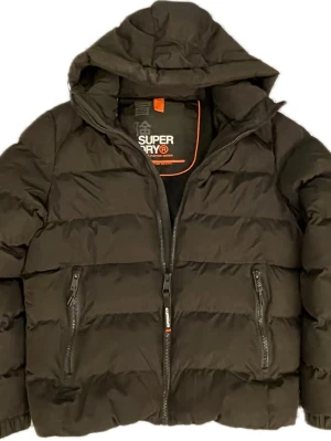Svart pufferjacka från Superdry - Säljer en svart pufferjacka från Superdry med huva och två dragkedjeförsedda fickor framtill. Jackan har en klassisk quiltad design och logotypdetaljer på bröstet och insidan. Perfekt för kalla dagar och riktigt skön att bära. Storlek L men sitter mer som M. Skick 9/10, använd endast ett fåtal gånger!😁