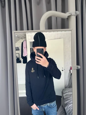 Svart hoodie från Morris - Snygg svart hoodie från Morris med guldfärgad logga på bröstet och justerbara snören i huvan. Klassisk passform och mjukt material som känns skönt mot huden. Perfekt för en avslappnad och stilren look.