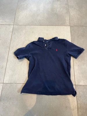 Marinblå pikétröja från Ralph Lauren - Klassisk marinblå pikétröja från Ralph Lauren med korta ärmar och krage. Tröjan har knäppning framtill och den ikoniska röda loggan broderad på bröstet. Perfekt för en stilren och avslappnad look.