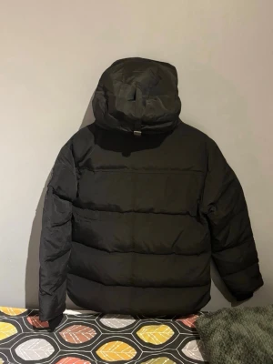 Svart pufferjacka från Canada Goose - Snygg svart pufferjacka från Canada Goose med huva och patch på ärmen. Jackan har en boxig passform, breda kanaler och dragsko i huvan. Perfekt för kalla dagar och riktigt trendig stil. Klassisk logga på bröstet och robust dragkedja framtill.