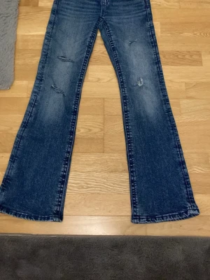Blå bootcut jeans med rosett-detalj för barn  - Säljer ett par blå bootcut jeans med slitningar på benen och snygga rosett-detaljer i silver på bakfickorna. Jeansen har en klassisk femficksmodell och är tillverkade i denim med lätt tvättad look. Perfekta för dig som gillar en retro och trendig stil.
