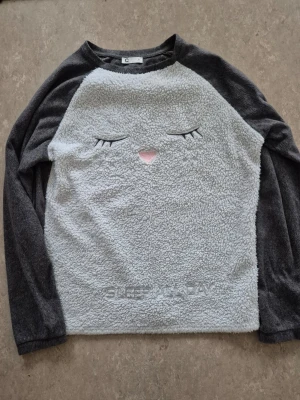 Tröja - Mysig tröja med fluffig ljusgrå framsida och mörkgrå ärmar och rygg. Framsidan har ett broderat sött ansikte med stängda ögon och rosa näsa samt texten 'SLEEP ALL DAY' längst ner. Perfekt för chill dagar hemma.