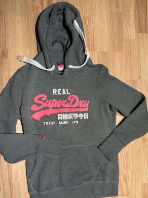 Grå Superdry hoodie med tryck - Säljer en nästan helt oanvänd unik superdry hoodie med fint rosa och vitt tryck framtill samt ribbade snören. Storlek S och sitter slim mot kroppen så väldigt bra passform men har inte kunnat ha på mig den då den inte passar och jag fick den😊 Bara att höra av er vid fler frågor!❤️