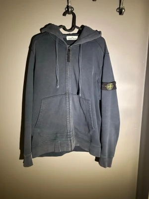 Mörkblå hoodie från Stone Island - Snygg mörkblå hoodie från Stone Island med dragkedja, huva och kängurufickor. Klassisk logotyp-patch på vänster ärm och justerbara snören i huvan. Perfekt för en avslappnad streetwear-look. Vänster armbåge är det slitage men inget som man tänker på