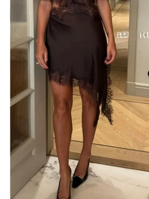 Brun asymmetrisk klänning med spets - Supersnygg svart kortklänning från H&M med asymmetrisk nederkant och spetsdetaljer längs kanterna. Klänningen har cut-out i midjan och cape-liknande överdel som ger en unik look. Perfekt för dig som vill sticka ut med en trendig och elegant stil.