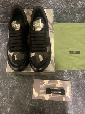 Valentino rockrunners camo sneakers i svart och grått - Snygga sneakers från Valentino med svart och grått camomönster, svarta skosnören och detaljer i mocka och läder. Skorna har en cool nitar-detalj vid hälen och en chunky sula som ger streetkänsla. Perfekta för dig som vill sticka ut med exklusiv design.