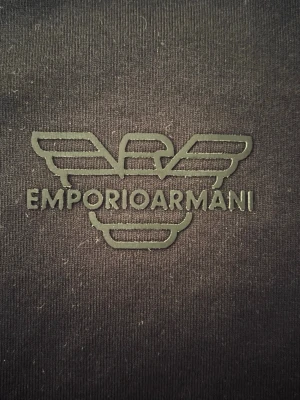 Svart Emporio Armani t-shirt - Snygg svart t-shirt från Emporio Armani med det klassiska logotrycket på bröstet. T-shirten har rund hals och korta ärmar, tillverkad i mjuk bomull för en skön känsla. Perfekt för dig som gillar stilrena och exklusiva plagg.