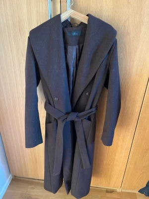 Svart lång kappa från Ellos - Stilren brun lång kappa från Ellos med bred krage och knytskärp i midjan. Kappan har dubbelknäppning och två stora fickor framtill. Perfekt för en trendig och klassisk look under kallare dagar.
