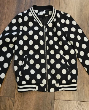 Svart bomberjacka med vita prickar - Snygg svart bomberjacka från Artigli med stora vita prickar och dragkedja framtill. Jackan har ribbade muddar med vita ränder vid ärmslut, krage och nederkant. Två fickor på sidorna och klassisk bomberjacke-look. Perfekt för dig som gillar statement-plagg.