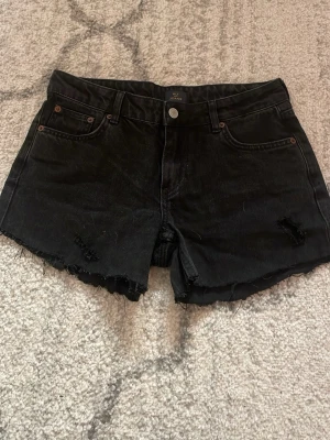 Svarta denimshorts från nelly - Snygga svarta denimshorts från Nelly med låg midja och råa kanter. Klassisk femficksmodell med knapp och dragkedja framtill. Lite slitna detaljer för en cool look. Perfekta för sommaren!