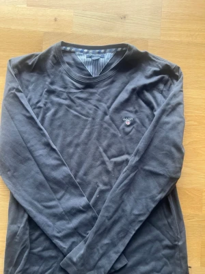 Mörkblå långärmad tröja från GANT - Snygg mörkblå långärmad tröja från GANT med rund halsringning och diskret logga broderad på bröstet. Insidan av kragen har ett blåvitt randigt tyg som ger en extra detalj. Perfekt till jeans eller chinos. Som ni ser på bröstkorgen så är de lite missfärgat osäker vad det är för något
