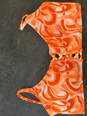 Orange och vit mönstrad bikini överdel  - Säljer en snygg bikini från Triumph med orange och vitt swirl-mönster. Bikinin har smala axelband och dekorativa detaljer mellan kuporna. Perfekt för strandhäng eller poolparty i sommar! Aldrig använd 