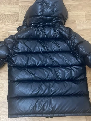 Svart pufferjacka med huva - Säljer en svart pufferjacka med glansig finish och stor huva. Jackan har två snedställda fickor med dragkedja och quiltad design som ger en riktigt puffig och trendig look. Perfekt för kalla vinterdagar och streetwear-stil. Materialet är syntet och jackan har dragkedja framtill.