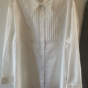 Vit skjorta med plisserad front från JTM - Klassisk vit skjorta från JTM med plisserad front och knappar hela vägen. Skjortan har lång ärm och en snygg krage som ger en stilren look. Perfekt att styla till olika outfits för en clean och trendig vibe.