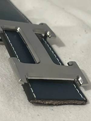 Mörkblått läderbälte med H-spänne - Snyggt mörkblått bälte i läder med kontrasterande vita sömmar och ett stort, silverfärgat H-format metallspänne. Perfekt accessoar för att lyfta din outfit med en stilren och modern touch. Kontakt MIG!! För flera bilder samt info,, den har två extra hål ja gjorde själv och klippte lite men fortfarande i mycket bra skick