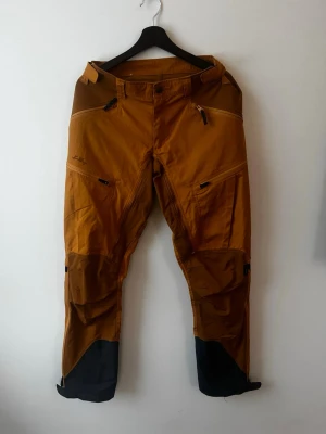 Lundhags jaktbyxor  - Säljer ett par gold dark/ gold Lundhags byxor med flera praktiska fickor, dragkedja, justerbar midja och är perfekta för utomhusaktiviteter. Materialet är slitstarkt och passar för tuffare väder. Använda väldigt lite så är som nya. 
