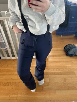 Mörkblå skidbyxor/termobyxor - Mörkblå skidbyxor med svarta justerbara hängslen och dragkedjeförsedda fickor framtill. Byxorna har bred passform och är tillverkade i ett slitstarkt, vattenavvisande syntetmaterial. Perfekta för vinterns alla äventyr i snön. 