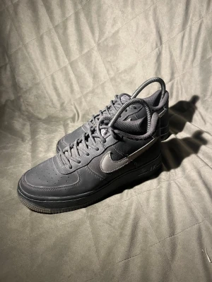 Nike Air Force 1 svarta sneakers - Säljer ett par klassiska Nike Air Force 1 sneakers i helsvart läder med svart sula och grå swoosh på sidan. Lågt skaft, perforerad tå och ikonisk AIR-detalj på mellansulan. Perfekta för dig som gillar stilrena och tidlösa sneakers.