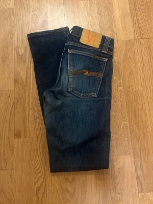 Mörkblå jeans från Nudie Jeans - Säljer ett par klassiska mörkblå jeans från Nudie Jeans med raka ben och snygga kontrastsömmar. Jeansen har en ikonisk bakficka med gul brodyr och patch i läderimitation i midjan. Perfekta för en avslappnad och stilren look.