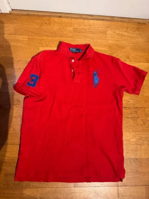 Röd pikétröja från Ralph Lauren - Klassisk röd pikétröja från Ralph Lauren med blå logga broderad på bröstet och siffran 3 på ärmen. Tröjan har krage, knappar framtill och är tillverkad i bomull. Perfekt för en sportig och stilren look. Sitter som M material är andvänt