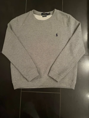 Grå sweatshirt från Polo Ralph Lauren - Klassisk grå sweatshirt från Polo Ralph Lauren i modellen Custom Fit. Tröjan har rund halsringning, långa ärmar och den ikoniska mörkblå logotypen broderad på bröstet. Tillverkad i mjukt bomullsmaterial för en skön och avslappnad look.
