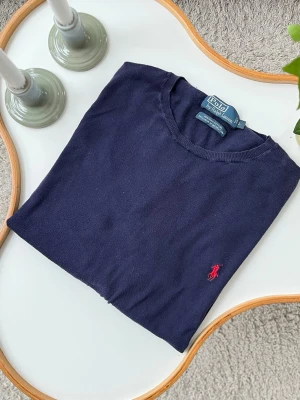 Mörkblå stickad tröja Polo Ralph Lauren i 100% pima bomull - Mörkblå stickad tröja från Polo Ralph Lauren med klassisk röd broderad logga på bröstet. Tröjan har rund halsringning, långa ärmar och ribbade muddar vid ärmslut och nederkant. Perfekt för lager på lager och stilren look. 100% pima bomull. Mycket bra skick, inga defekter förutom lappen är lös i ett hörn (väldigt lätt lagning). Storlek L. Axelbredd: 51 cm Längd: 66 cm