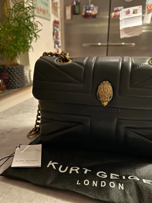 Svart quiltad handväska Kurt Geiger - Snygg svart quiltad handväska från Kurt Geiger med guldfärgad kedjerem och ikonisk örndekor på locket. Väskan har en mjuk struktur och detaljer i antikguld, perfekt för att lyfta vilken outfit som helst. Inga defekter, perfekt storlek 