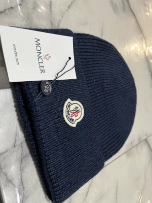 Mörkblå ribbstickad Moncler mössa - Snygg mörkblå ribbstickad mössa från Moncler med klassisk uppvikt kant och broderad logotyp framtill. Tillverkad i mjukt material som håller dig varm och ger en clean look. Perfekt accessoar för kyliga dagar.