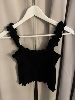Svart croppad linnetopp med volang - Svart croppad linnetopp med volangdetaljer längs axelbanden och nederkanten. Toppen har breda axelband och knappt använd från shein i storlek M