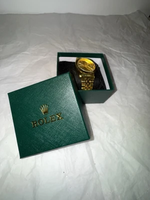 Guldfärgad klocka i box - Säljer en stilren guldfärgad klocka med klassisk länkarmband och rund urtavla. Klockan levereras i original grön box med guldfärgad logga. Perfekt accessoar för dig som vill sticka ut med lyxig känsla.