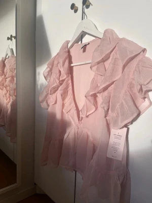 Ljusrosa volangblus från Nelly - Supersöt ljusrosa blus från Nelly med djup V-ringning och stora volanger längs axlar och bröst. Tillverkad i ett tunt, transparent tyg som ger en luftig känsla. Perfekt för dig som gillar romantisk och trendig stil.