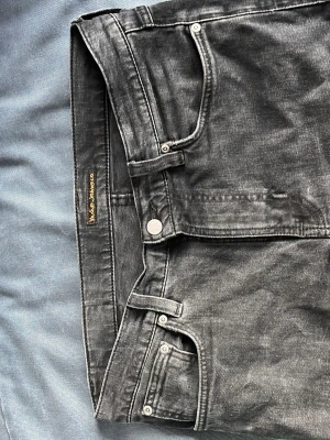 Svarta jeans från Nudie Jeans Co - Snygga svarta jeans från Nudie Jeans Co med klassisk femficksdesign och diskret logga på bakfickan. Jeansen har en tvättad look och raka ben. Perfekta för dig som gillar stilrena och tidlösa plagg i garderoben.
