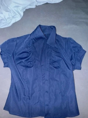 Mörkblå kortärmad skjorta - Snygg mörkblå skjorta med korta ärmar och klassisk krage. Skjortan har knappar framtill och lätt puffade detaljer vid axlarna. En lätt urringning vid halsen