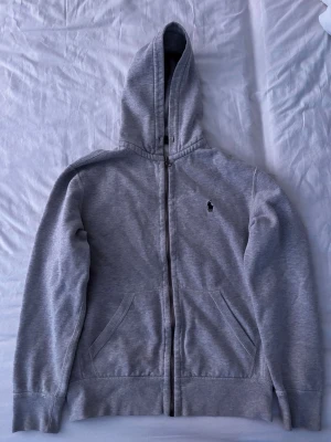Grå zip hoodie från Polo Ralph Lauren - Orginal pris: 1100-1300kr. Klassisk grå hoodie med dragkedja från Polo Ralph Lauren. Tröjan har huva, två fickor framtill och den ikoniska lilla broderade loggan på bröstet. Tillverkad i mjuk bomullsblandning som är skön att ha på sig. Perfekt för en avslappnad och stilren look. Två mini mini fläckar fram och dragkedjan är lite sliten.
