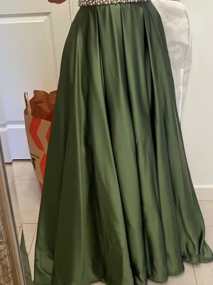 Grön långklänning med pärldetaljer - Säljer en elegant långklänning i mörkgrönt satinliknande material. Klänningen har en markerad midja med pärldetaljer och ett svepande, utsvängt kjolparti. Perfekt för bal eller speciella tillfällen.