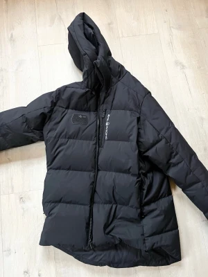 Svart dunjacka från Peak Performance - Säljer en svart dunjacka från Peak Performance med huva och dragkedja framtill. Jackan har en quiltad design, diskret logga på bröstet och är perfekt för kalla vinterdagar. Materialet är tåligt och håller värmen riktigt bra.