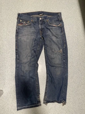 Blå slitna jeans från True Religion - Säljer ett par blå jeans från True Religion med slitningar och hål på benen för en cool, avslappnad look. Jeansen har raka ben, klassiska fem fickor och tydliga sömmar samt broderade bakfickor med röd detalj. Materialet är denim i bomull.