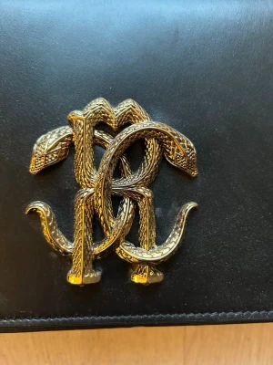 Svart axelväska från Roberto Cavalli - Köpt från Maruschka De Margo, där jag betalade 17.000kr  Snygg svart axelväska i slätt läder från Roberto Cavalli. Väskan har en guldfärgad kedjerem och ett stort, detaljerat ormformat RC-monogram i metall på framsidan. Perfekt statement piece för dig som gillar lyxiga accessoarer.