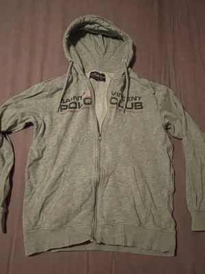 Grå hoodie Saint Vincent Polo Club - Grå hoodie med dragkedja från Saint Vincent Polo Club. Hoodien har tryck framtill, huva med dragsko och två fickor. Perfekt för en avslappnad och sportig stil.