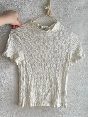 Vit topp från & Other Stories - Superfin vit topp från & Other Stories med korta ärmar och ett unikt stickat mönster. Toppen har volangdetaljer vid hals, ärmslut och nederkant som ger en söt touch. Perfekt att styla med jeans eller kjol för en trendig look.
