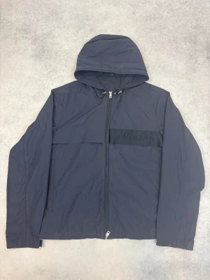 Svart vindjacka från Moncler - Stilren svart vindjacka från Moncler med huva och dragkedja framtill. Jackan har diskret Moncler-logga på bröstet och elastiska muddar vid ärmsluten. Tillverkad i lätt och tåligt material som passar perfekt för blåsiga dagar.