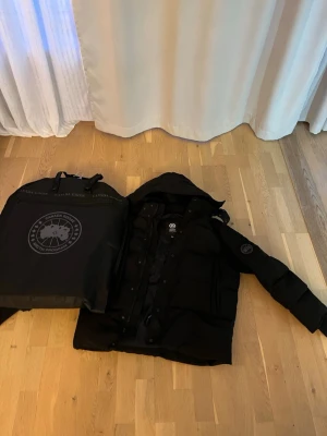 Svart dunjacka från Canada Goose - Sänk kan sänkas vid snabb affärSäljer en svart dunjacka från Canada Goose med huva och stora framfickor. Jackan har en klassisk patch på ärmen och stängs med både dragkedja och knappar. 