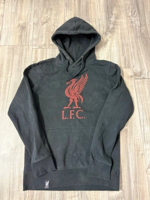 Svart Liverpool FC hoodie med tryck - Svart hoodie från Liverpool FC med stor röd logga och texten L.F.C. på bröstet. Klassisk känguruficka framtill och justerbar huva med snören. Tillverkad i mjukt bomullsmaterial, perfekt för dig som vill visa ditt stöd för laget.