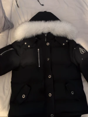 Svart moose knuckels jacka  - Snygg svart dunjacka från Moose Knuckles med avtagbar huva och vit pälskant. Jackan har silverfärgade dragkedjor, tryckknappar och logga på ärmen. Flera fickor framtill och en modern, puffig passform som håller dig varm under kalla dagar. L men sitter som M