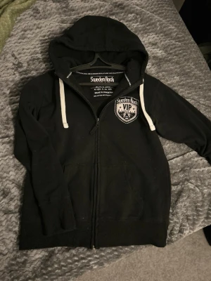 Svart Sweden Rock hoodie med dragkedja - Säljer en svart hoodie från Sweden Rock med vit logga och text på bröstet. Hoodien har dragkedja, vita snören i huvan och två fickor framtill. Tillverkad i 80% bomull och 20% polyester. Perfekt för dig som gillar rock och festivalstil.