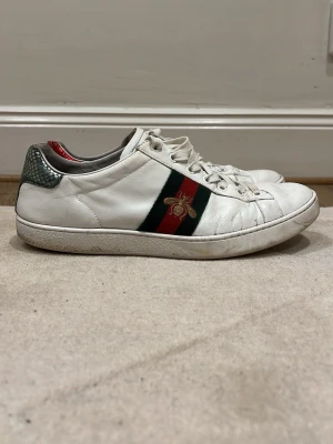 Gucci vita sneakers med bi-detalj - Snygga vita sneakers från Gucci med klassiska gröna och röda ränder på sidorna och en broderad guldfärgad bi. Hälkappan har metallicdetaljer i grönt och rött ormskinnsmönster. Skorna är i läder och har vita snören och sula.