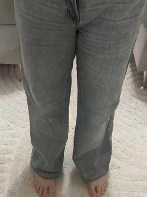 Grå raka jeans med hög midja - Säljer ett par grå jeans med rak passform och hög midja. Jeansen har klassiska fem fickor och stängs med dragkedja och knapp. Materialet är denim i bomull och färgen är tvättad grå, perfekt för en avslappnad stil.