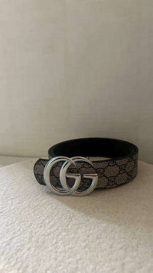  beige bälte med silverdetalj - Snyggt bälte från Gu€€i i beige och svart med klassiskt GG-mönster och stor silverfärgad GG-spänne framtill. Bältet har en bred design och är perfekt för att lyfta vilken outfit som helst. ej äkta! Går att använda. Dålig kvalitet 