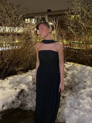 Svart långklänning med öppen rygg - Elegant svart långklänning med axelbandslös design och öppen rygg. Klänningen har en snygg scarf-detalj som faller längs ryggen och en figurnära passform upptill. Perfekt för dig som vill sticka ut med en stilren look.